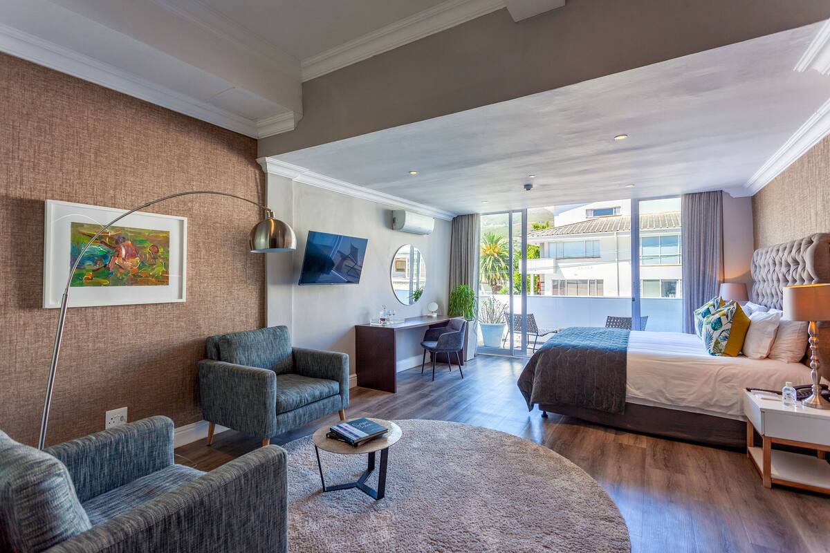 A modern hotel room with a large bed, a desk, a chair, and a sofa. There is a large window providing a view of the outside.