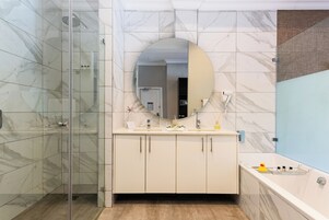 A bathroom with a glass shower enclosure, a marble-tiled wall, a round mirror, a white vanity with two sinks, and a bathtub with a rubber duck.