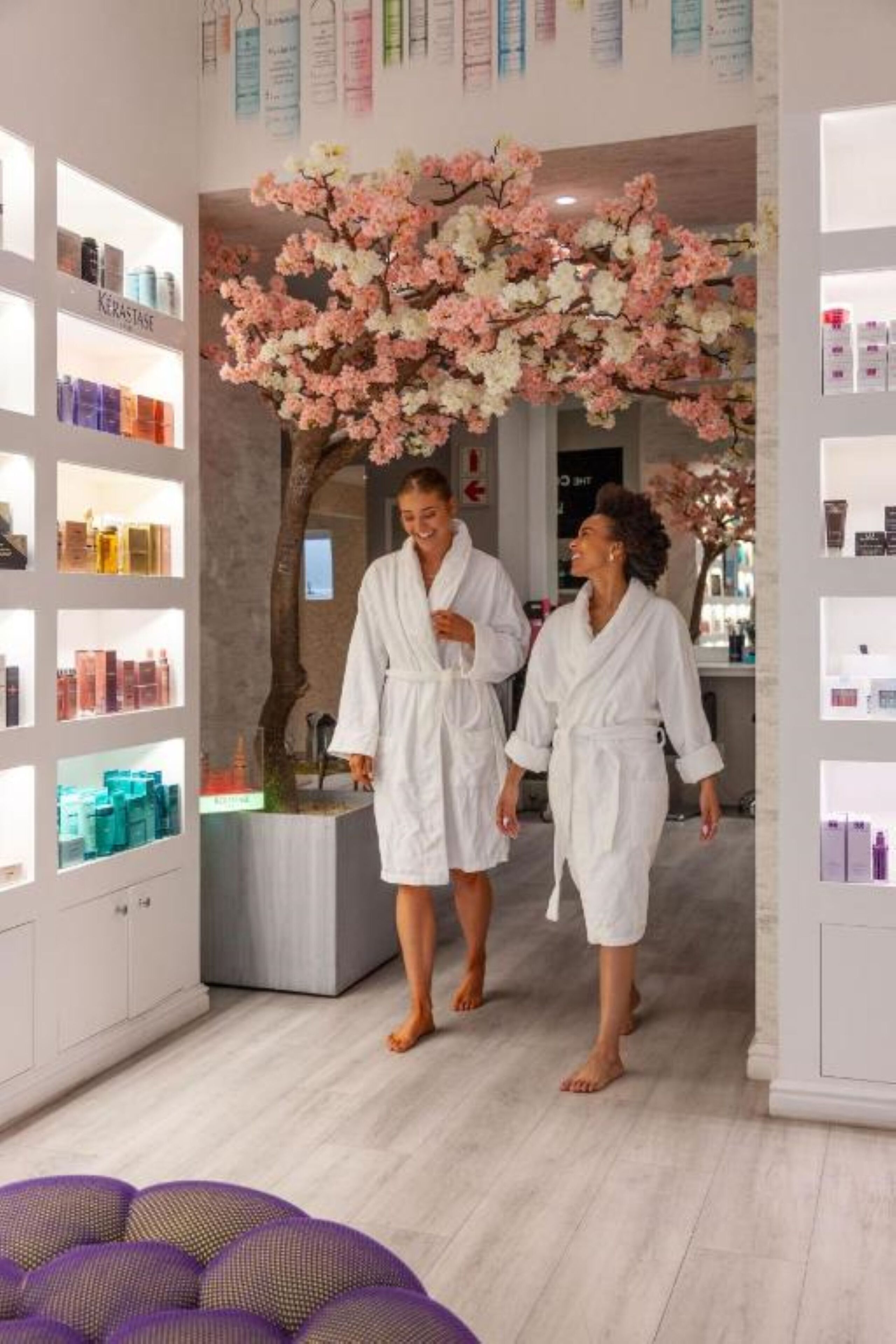 A spa area with shelves of skincare products, a large artificial cherry blossom tree, and two people in white robes.