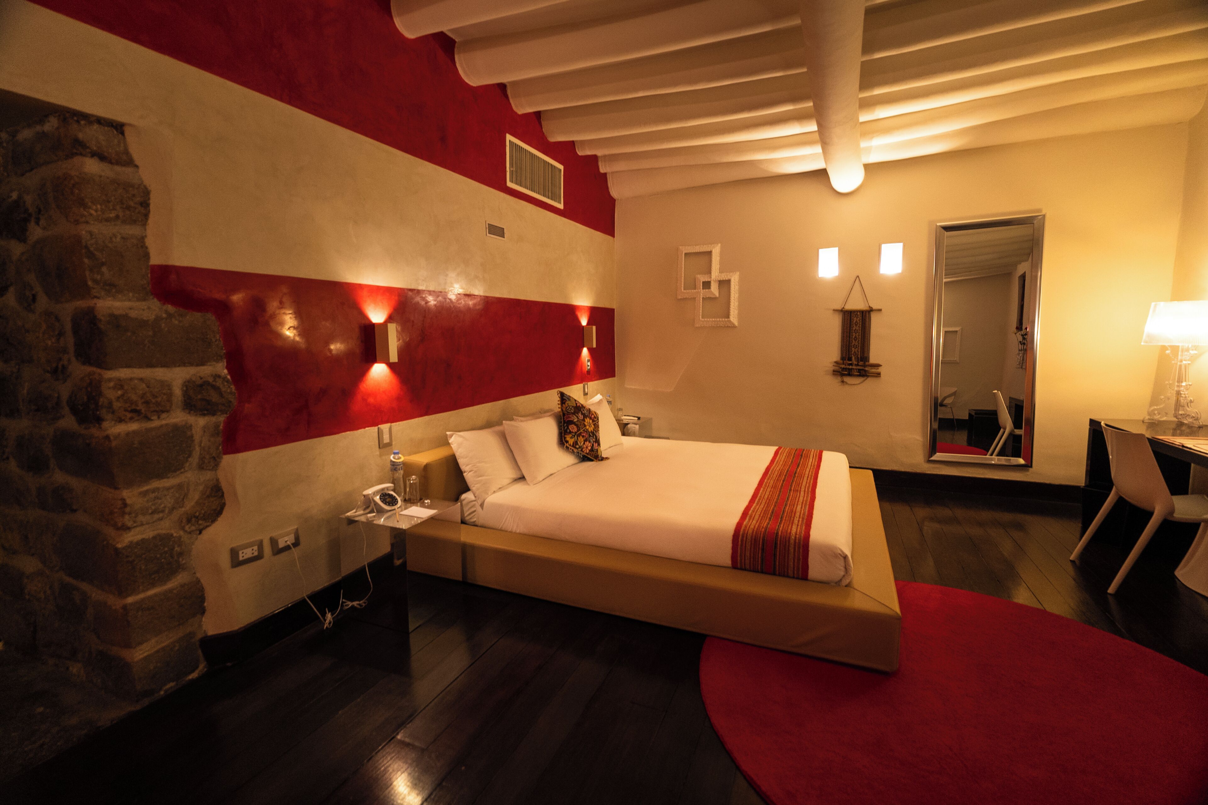 A hotel room with a bed, a stone wall, a red and white wall, a desk, and a chair.