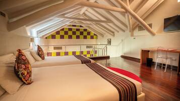 A spacious room with a high ceiling, a checkered wall, a bed with a striped blanket, a desk with a chair, and a TV mounted on the wall.