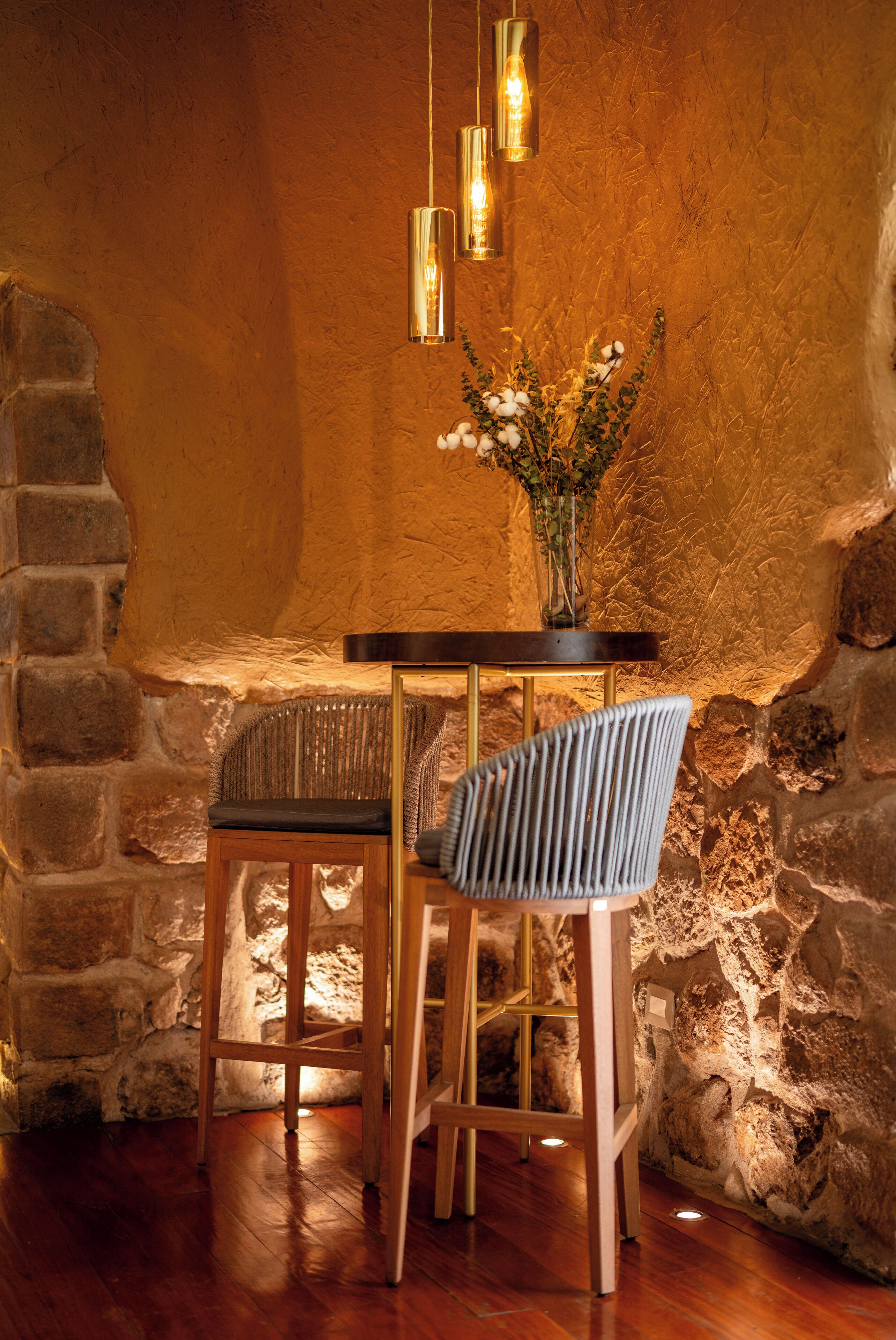 Un intérieur rustique avec un mur en pierre, une table en bois ornée d’un vase de fleurs et deux chaises à dossier en osier.