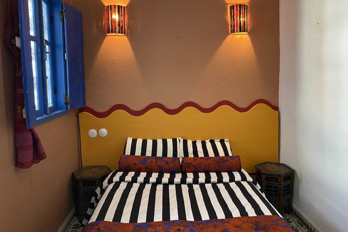 Un dormitorio con cubrecama a rayas, dos lámparas fijadas a la pared, un ribete rojo ondulado y una puerta azul.