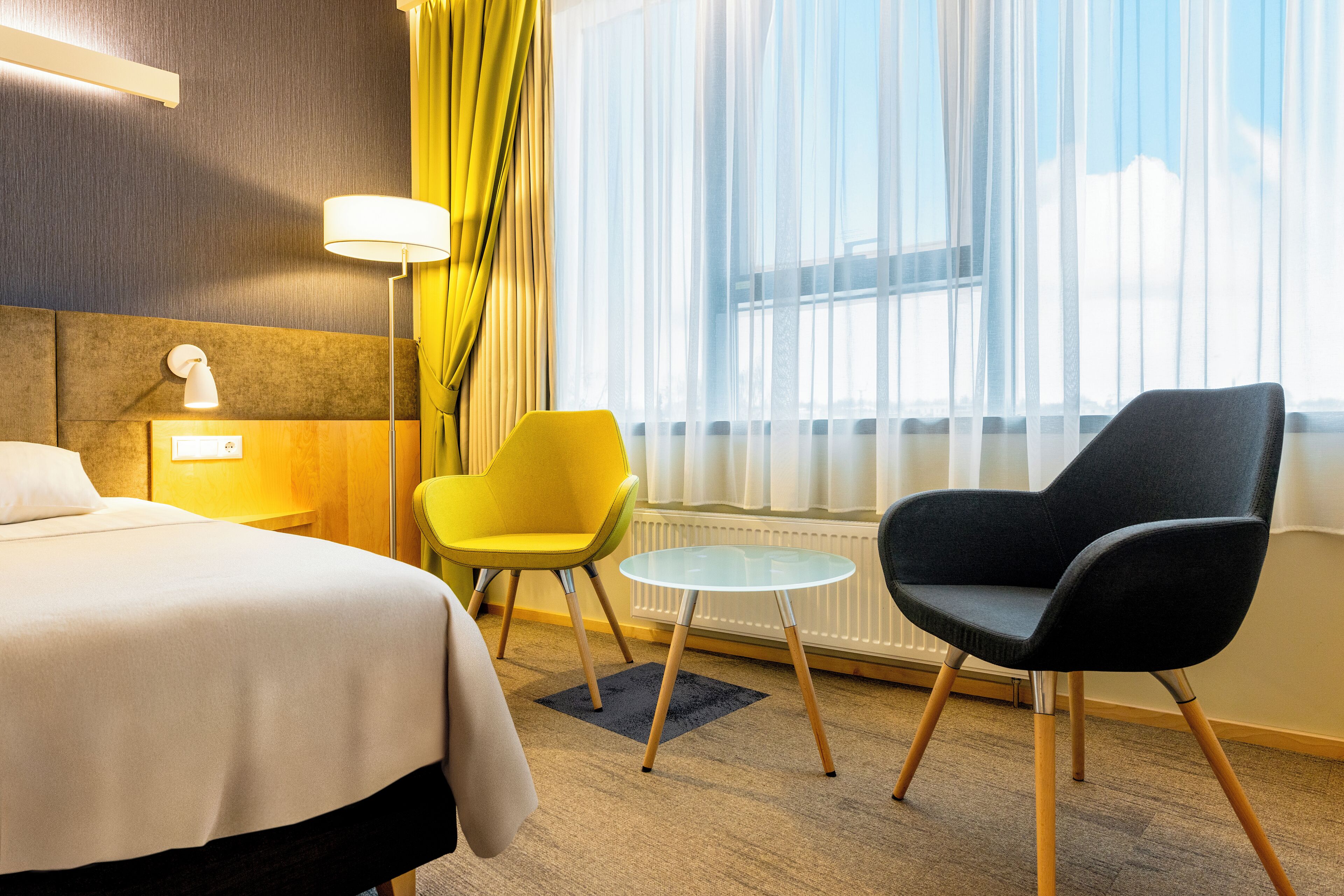 Habitación de hotel con cama, una silla amarilla, una silla negra, una mesita redonda y una ventana con cortinas traslúcidas.