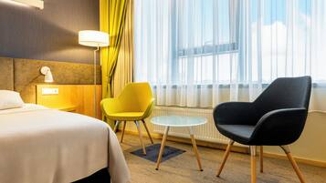 Habitación de hotel con cama, una silla amarilla, una silla negra, una mesita redonda y una ventana con cortinas traslúcidas.