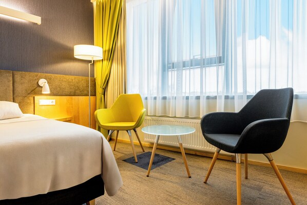 A hotel room with a bed, a yellow chair, a black chair, a small round table, and a window with sheer curtains. - Tartu Hotel (Tartu)