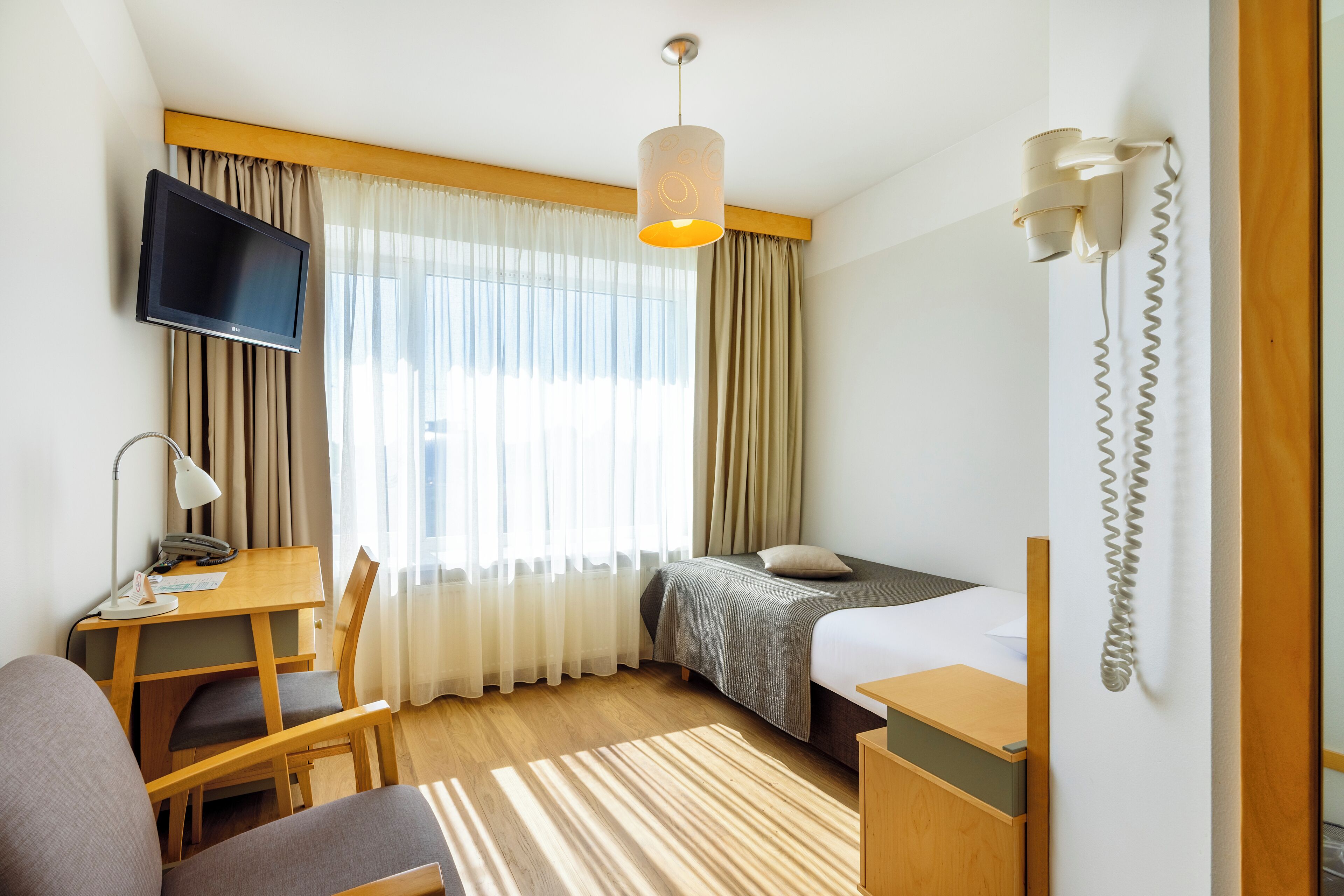A hotel room with a bed, a desk with a chair, a TV mounted on the wall, and a telephone.