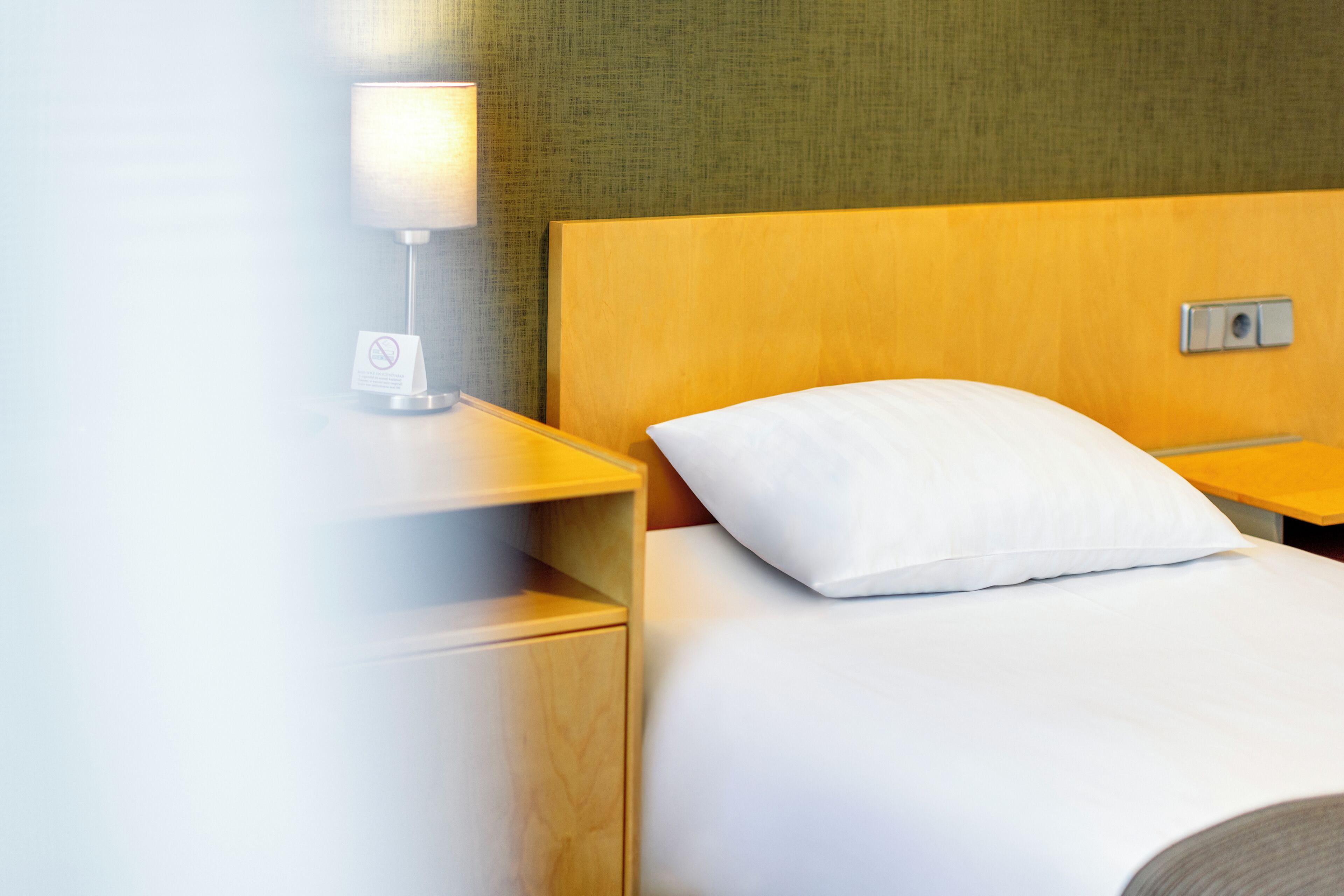 Habitación de hotel con cabecera de madera, almohada blanca, mesita de noche con lámpara e interruptor de luz en la pared.