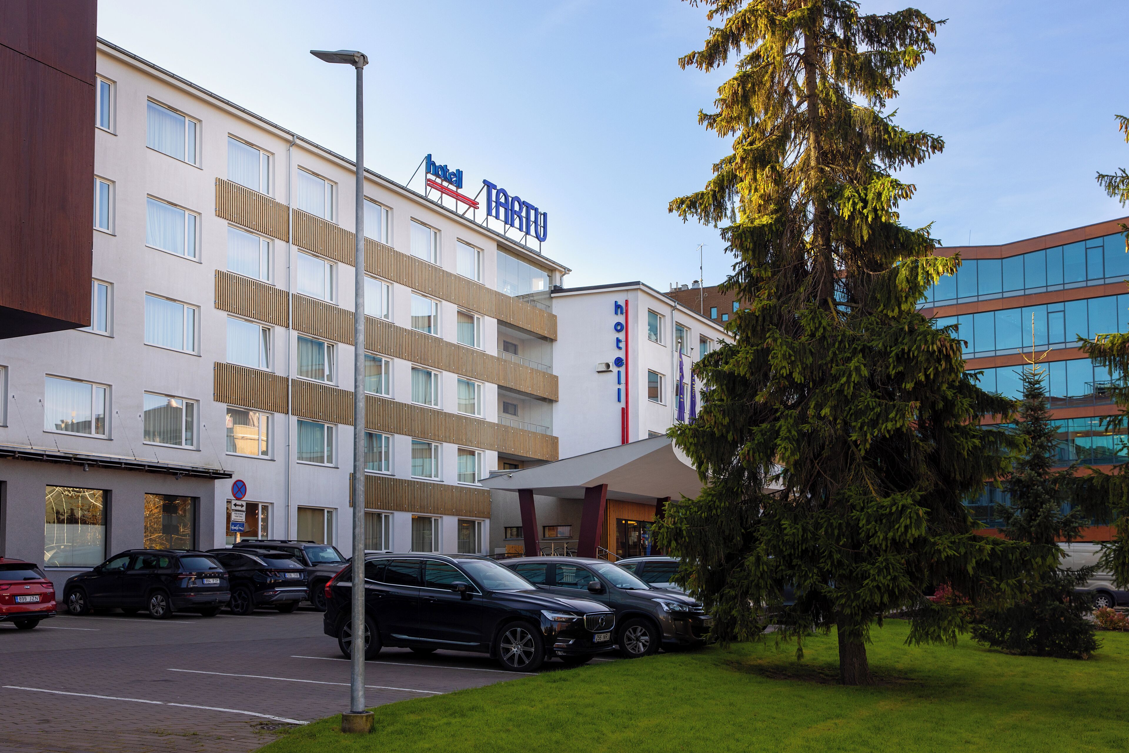 Un hotel moderno de varios pisos, un estacionamiento con varios coches y un árbol grande en primer plano.