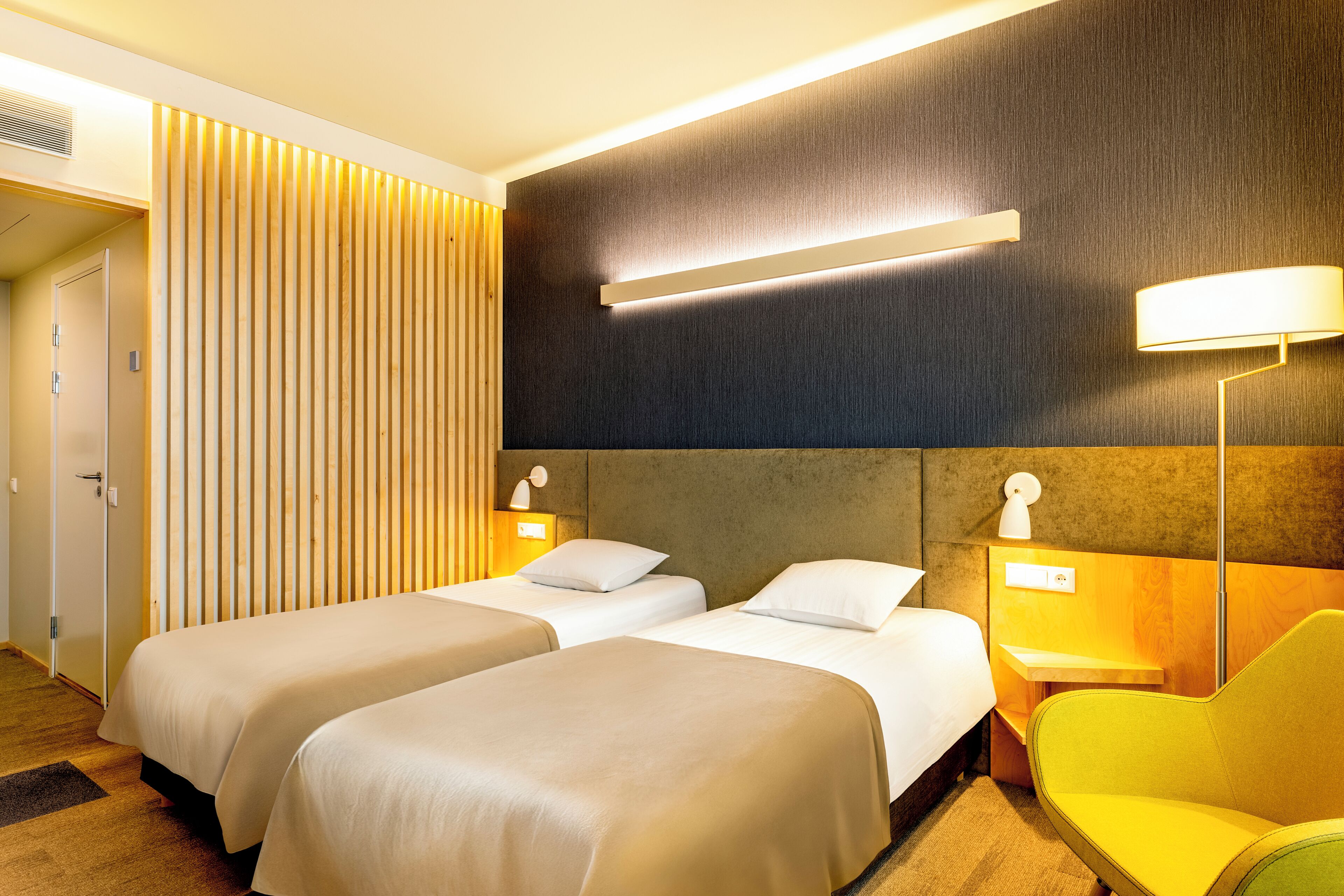 A hotel room with two beds, a wooden slat wall, a modern lamp, and a yellow chair.