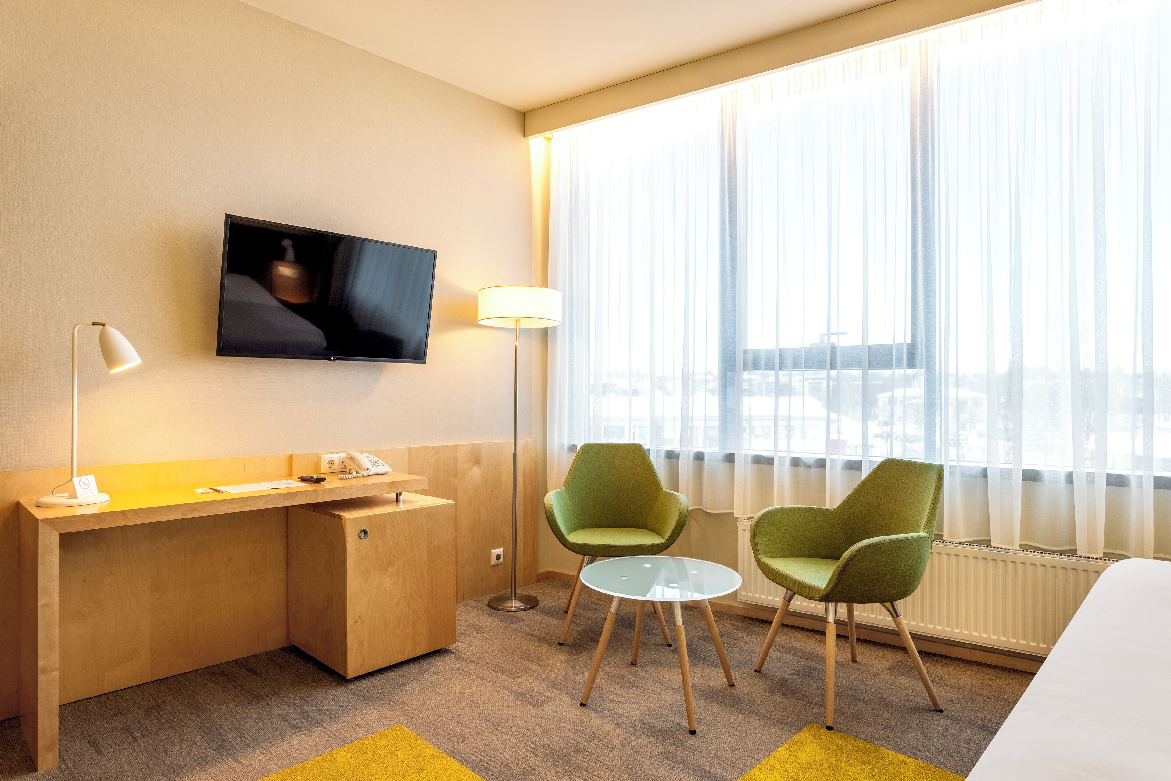 A hotel room with a wooden desk, two green chairs, a television, and a window with sheer curtains.