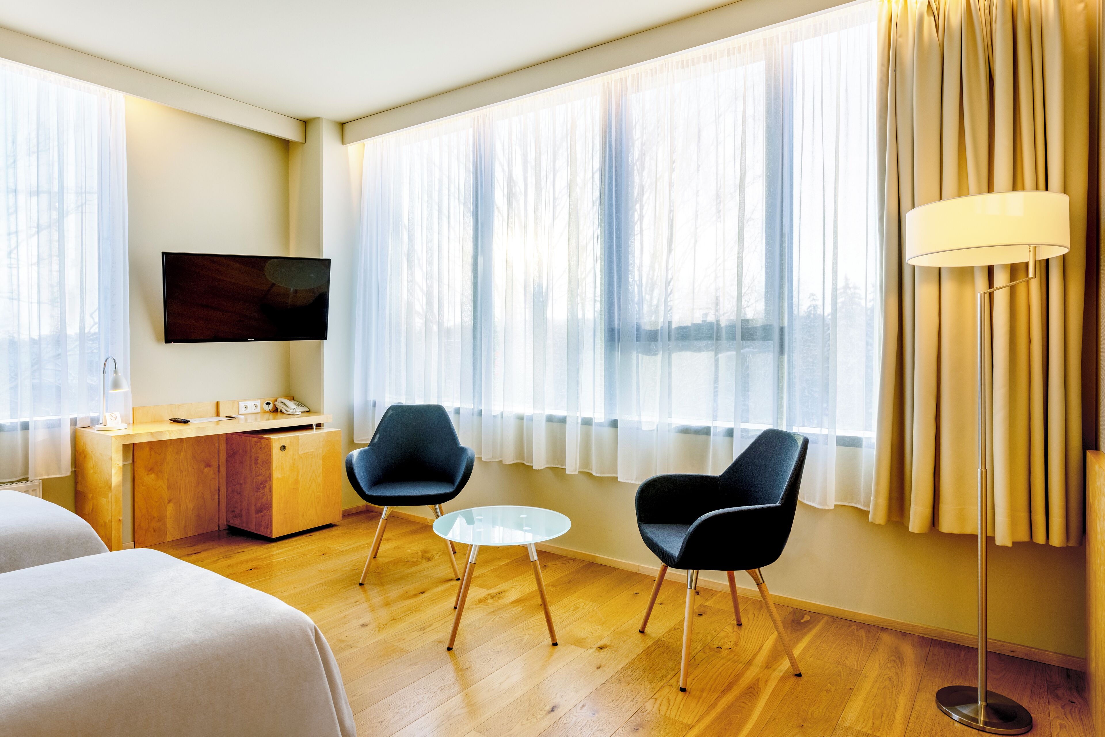Une chambre d’hôtel avec un bureau en bois, deux chaises noires, une petite table ronde, un téléviseur fixé au mur et de grandes fenêtres avec des rideaux légers.
