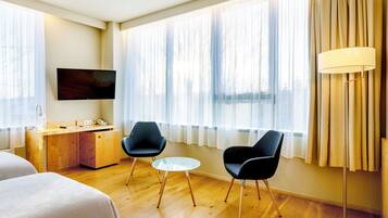 Une chambre d’hôtel avec un bureau en bois, deux chaises noires, une petite table ronde, un téléviseur fixé au mur et de grandes fenêtres avec des rideaux légers.