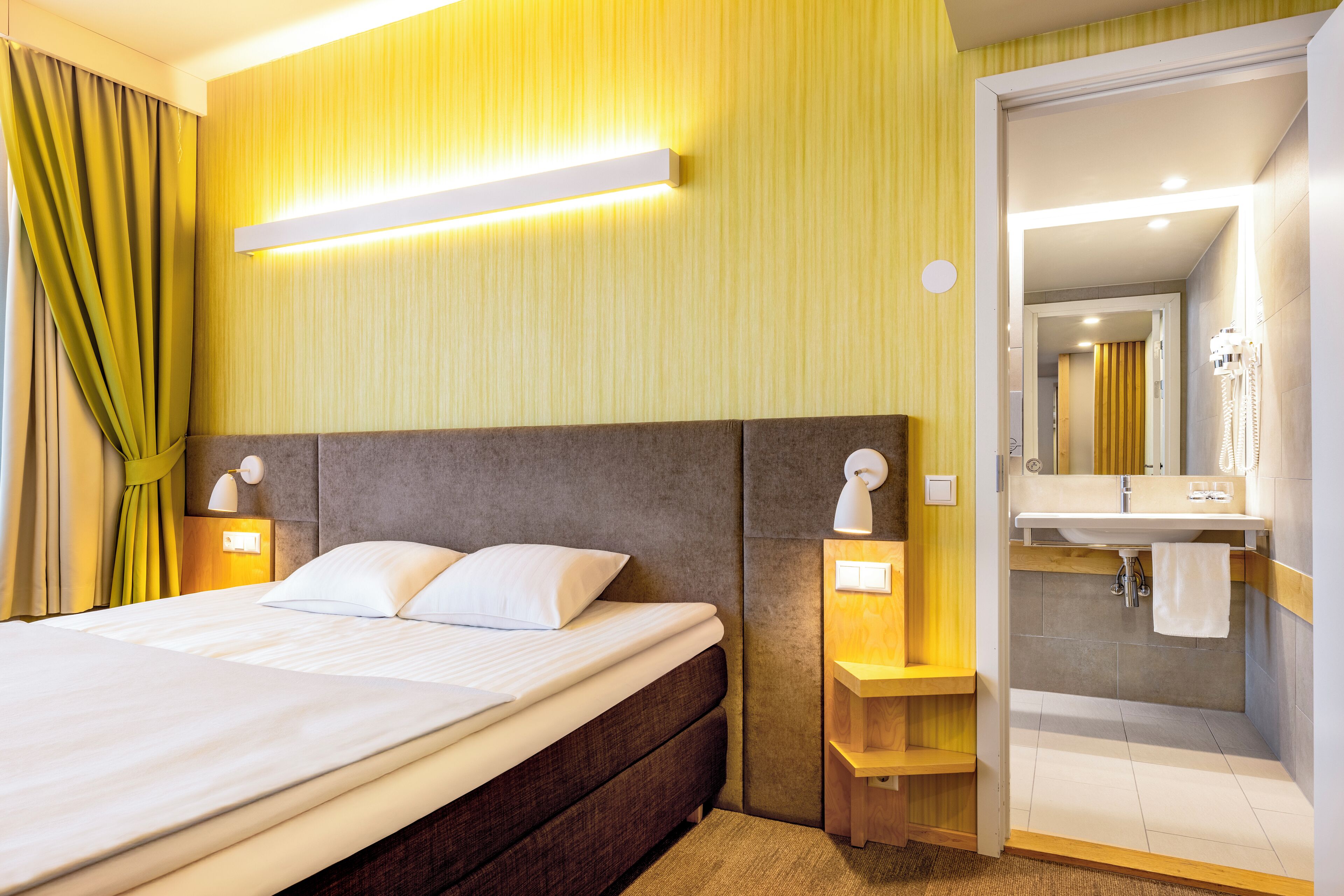 A hotel room with a bed, bedside lamps, a nightstand, a mirror, and a bathroom with a sink and towel rack.