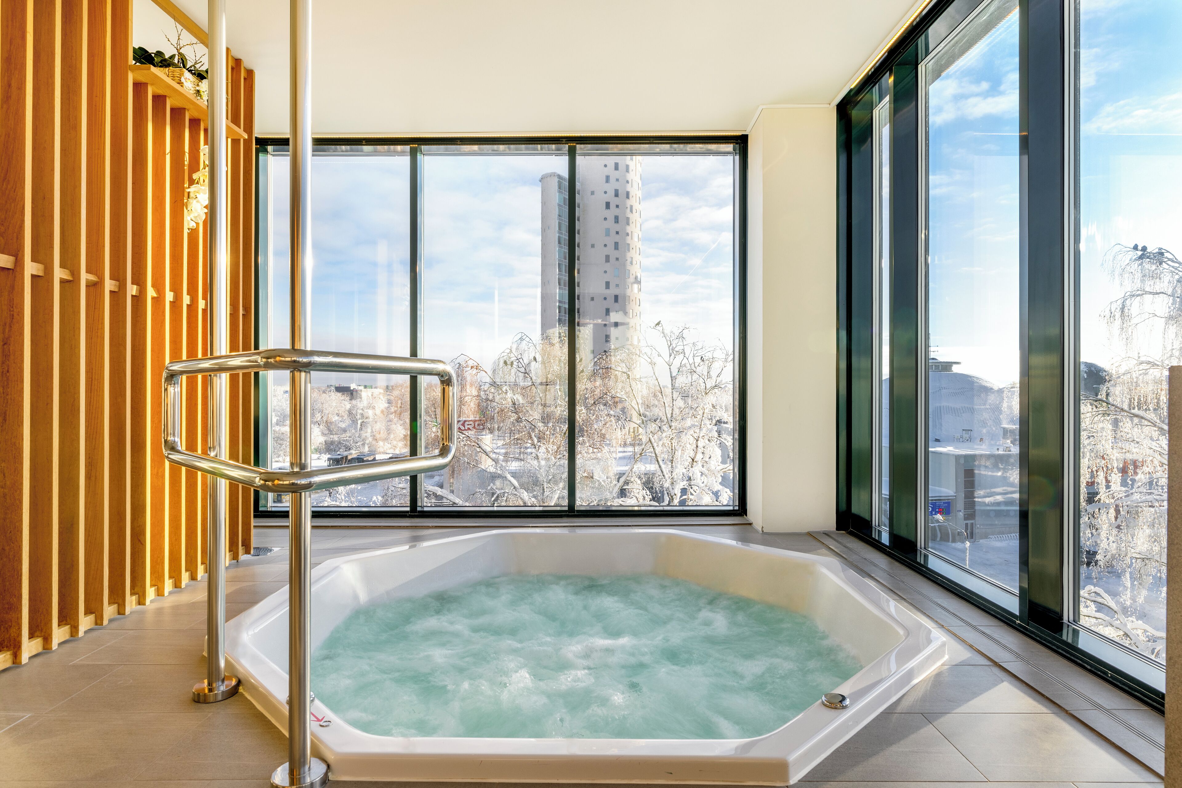 A modern bathroom with a large bathtub, a wooden panel wall, and a city view through large windows.