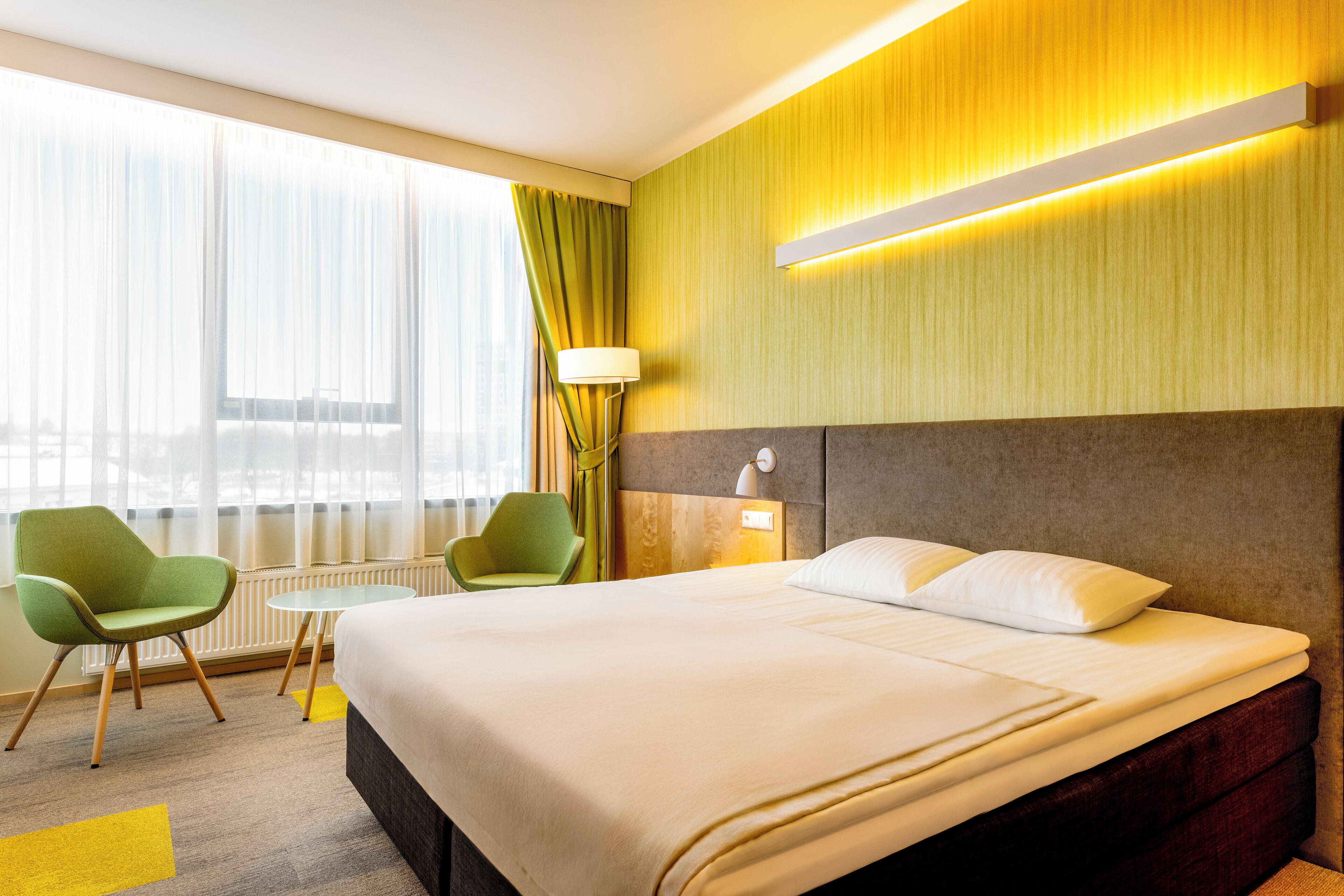 A hotel room with a large bed, two green chairs, a small table, and a window with sheer curtains.