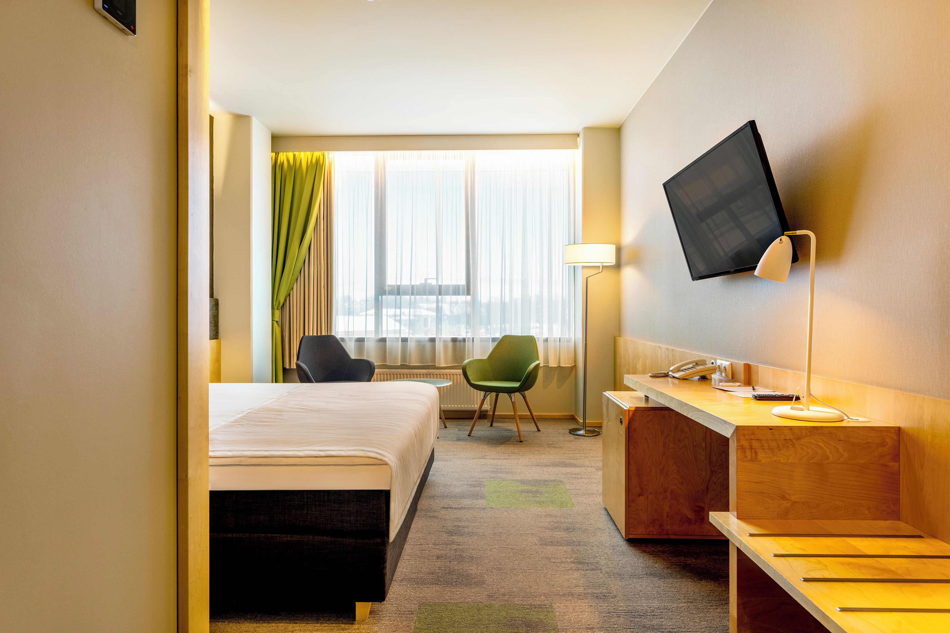 Una habitación de hotel con cama, escritorio, silla, televisión y ventana con cortinas.
