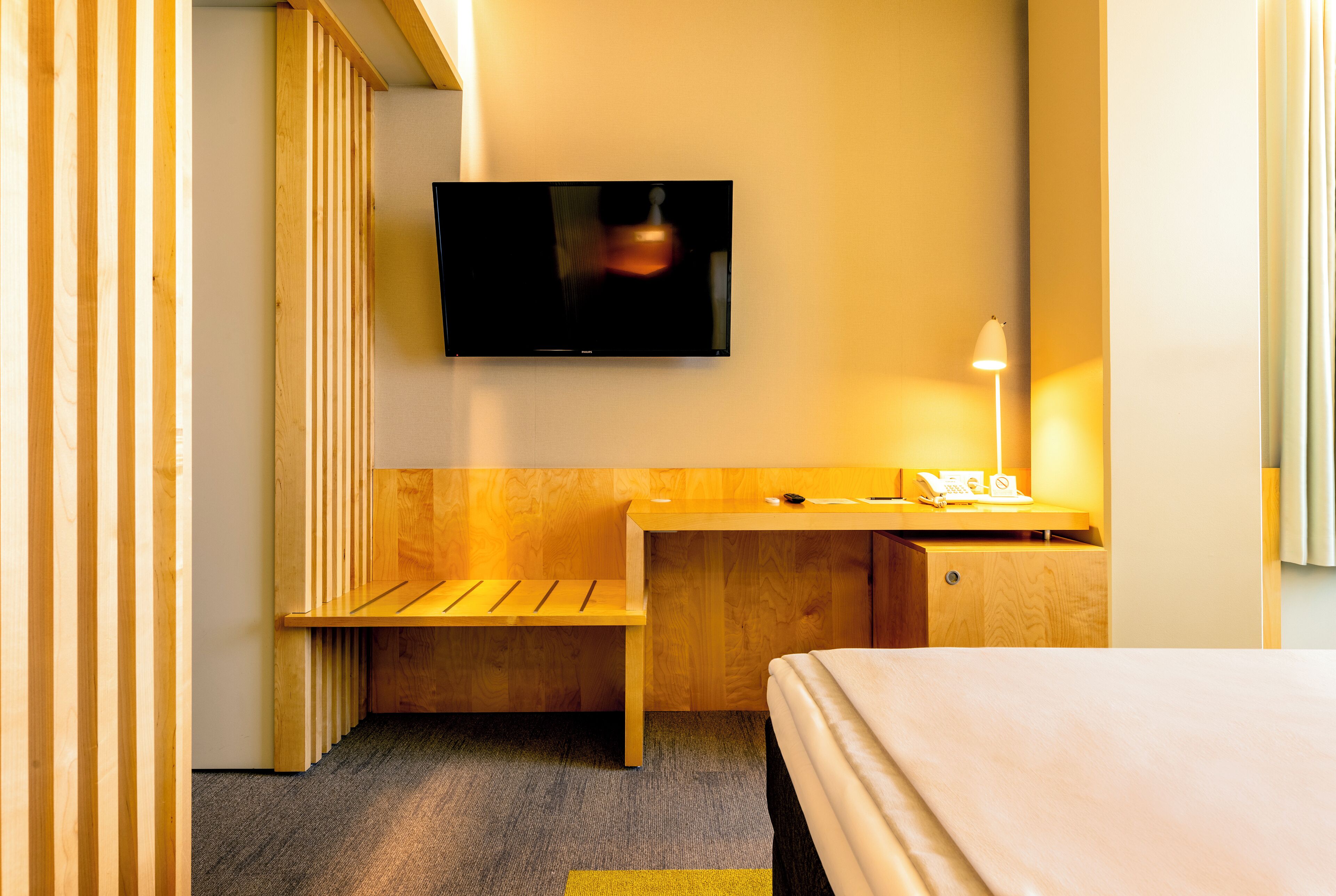 A hotel room with a bed, a desk with a lamp, a television, and a wooden bench.