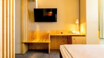 A hotel room with a bed, a desk with a lamp, a television, and a wooden bench.