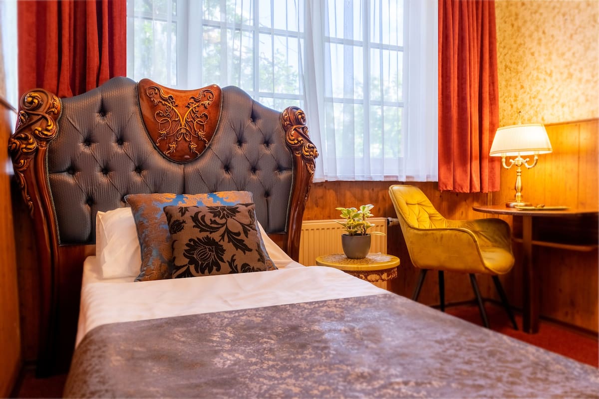 A bedroom with a tufted headboard, a bed with white and patterned pillows, a small round table with a lamp, a chair, and a potted plant.