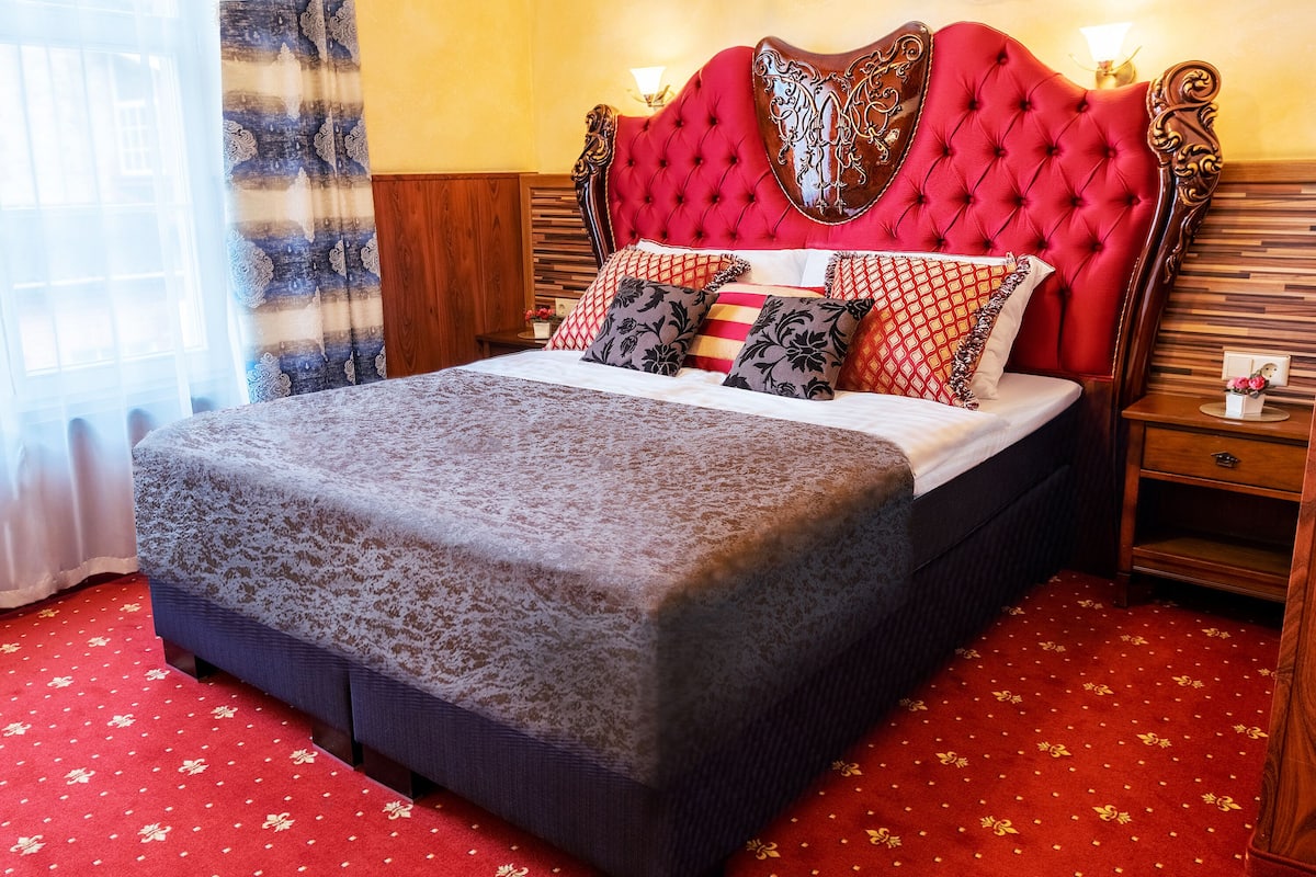 A large bed with a red headboard, multiple pillows, and a wooden nightstand with a lamp.