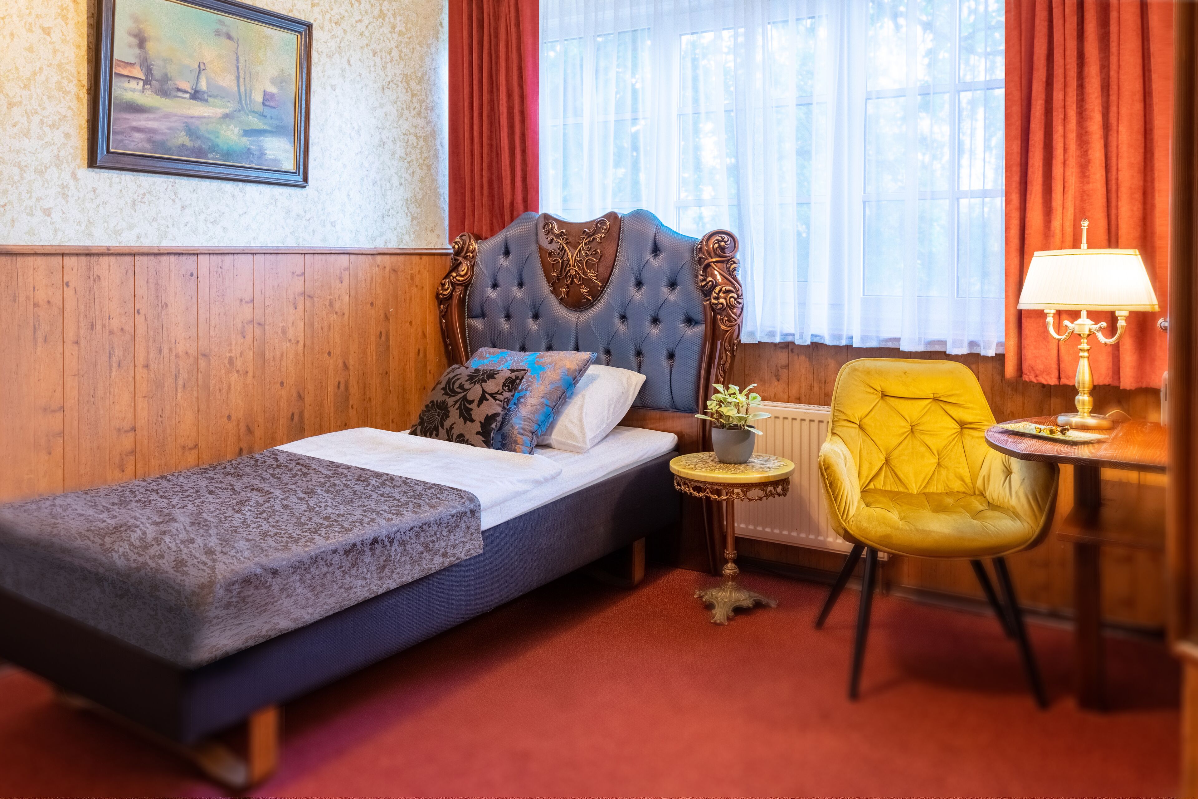Een eenpersoonsbed met hoofdbord, een klein tafeltje met lamp, een gele fauteuil en een schilderij aan de muur.