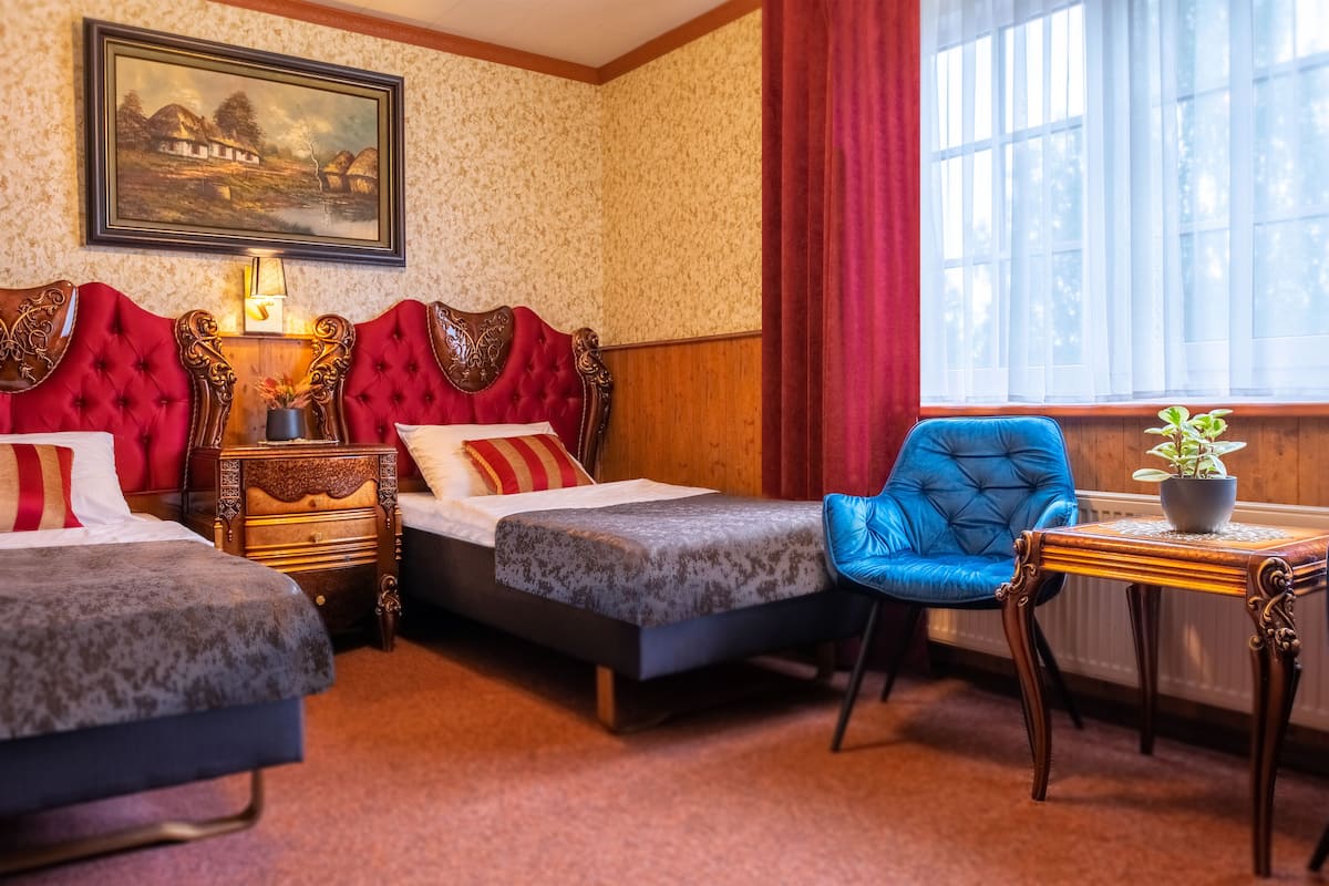 A hotel room with two beds, a blue chair, a small table with a plant, and a painting on the wall.