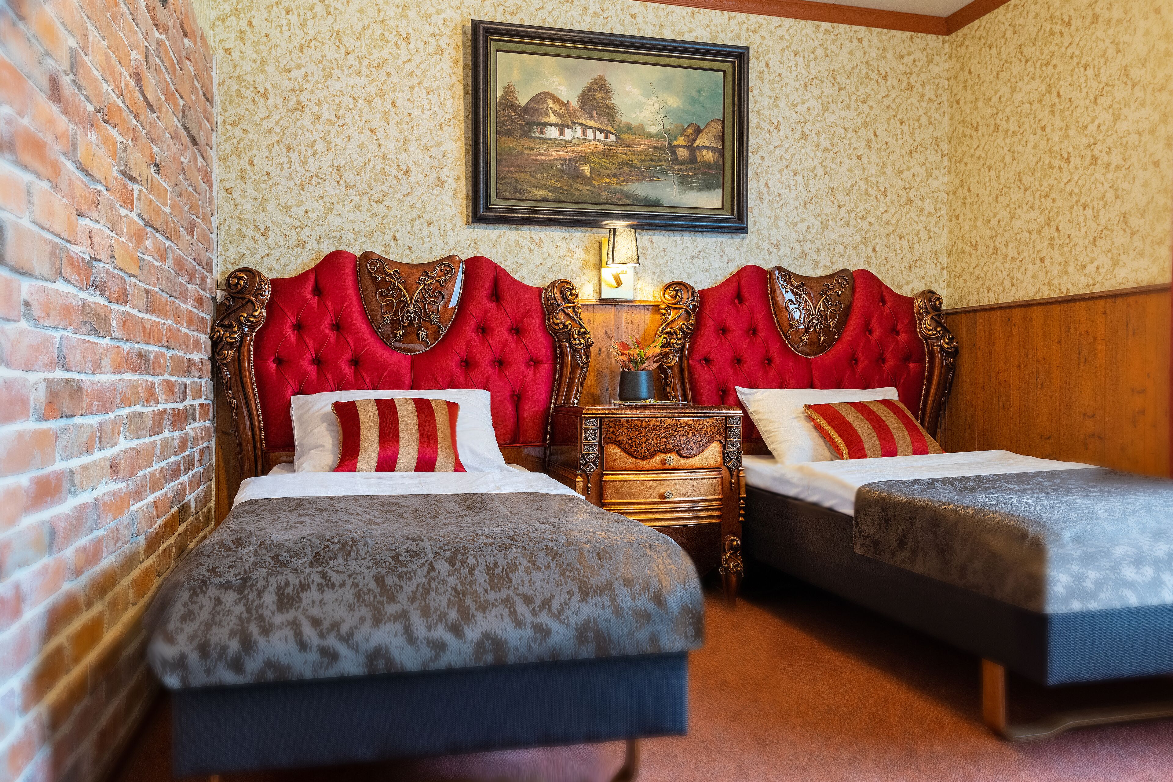 Een kamer met twee bedden, een bakstenen muur, een houten hoofdbord, een schilderij en een nachtkastje met een lamp.