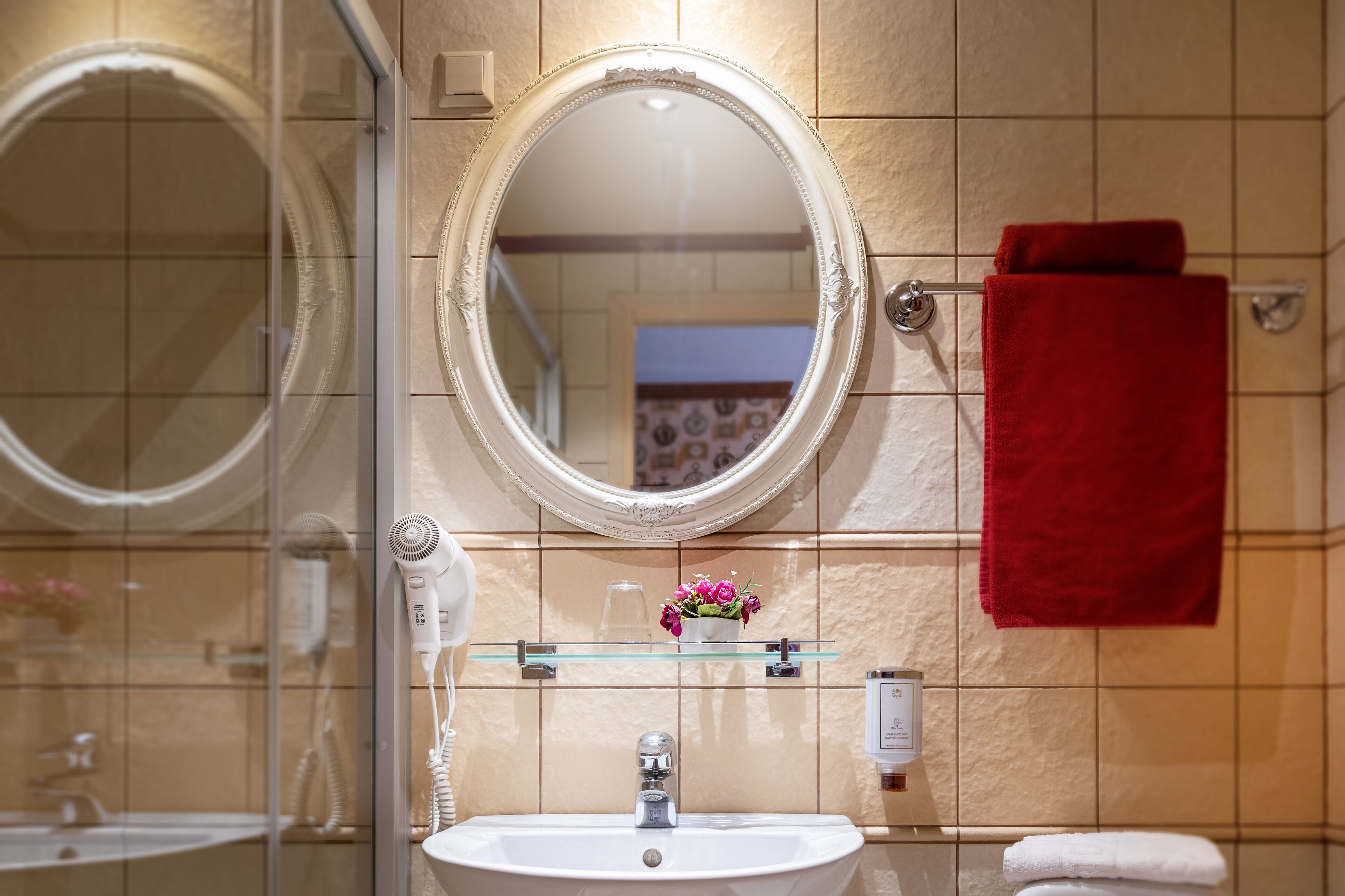 A bathroom with a white sink, a glass shelf with a vase of flowers, a red towel, and a large mirror.