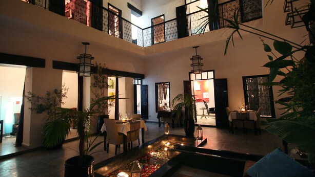 A spacious indoor area with multiple dining tables and chairs, a central water feature with candles, and a balcony with decorative railings. - Riad Villa Wengé (Marrakech)