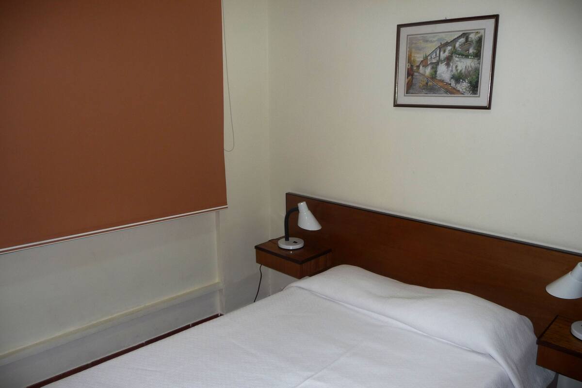 Siisti hotellihuone, jossa on huolitellusti pedattu sänky, yöpöytä lampun kanssa, seinällä oleva kehystetty kuva ja ikkunassa sälekaihtimet.