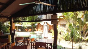 A covered outdoor seating area with wooden tables and chairs, a pool, and tropical plants. - Trees Too Guest Lodge (Nkomazi)