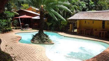 Relax by the serene pool surrounded by lush greenery and a thatched-roof structure.