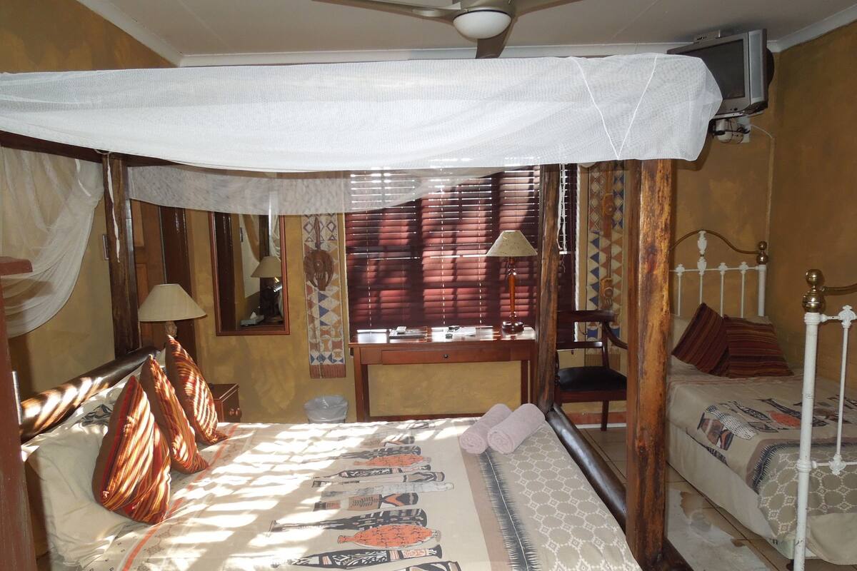 Quarto com cama, escrivaninha, cadeira, televisão e ventoinha no teto.