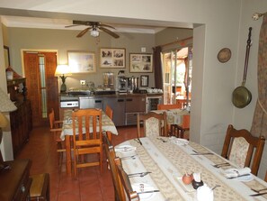Un'area pranzo con un tavolo lungo apparecchiato per quattro persone, sedie, una ventola a soffitto e una cucina con elettrodomestici e una macchina da caffè.