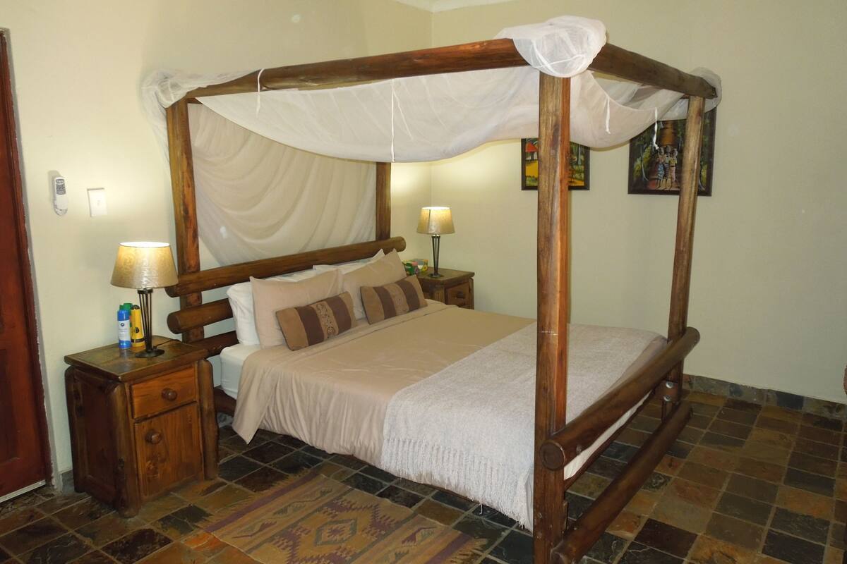 Uma cama de dossel com cortina, mesas de cabeceira de madeira, um tapete estampado e um quadro na parede.
