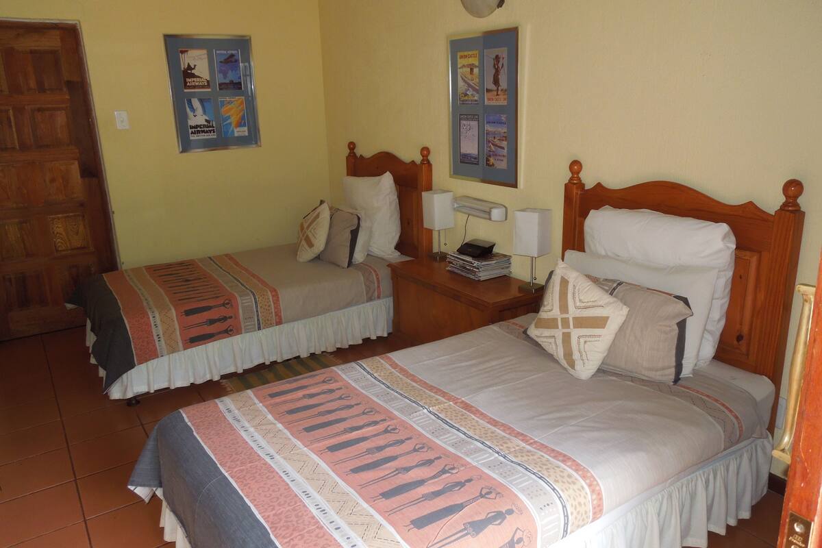 Quarto com duas camas, cada uma com cabeceira de madeira, mesa de cabeceira com abajur e um cartaz fixado na parede.