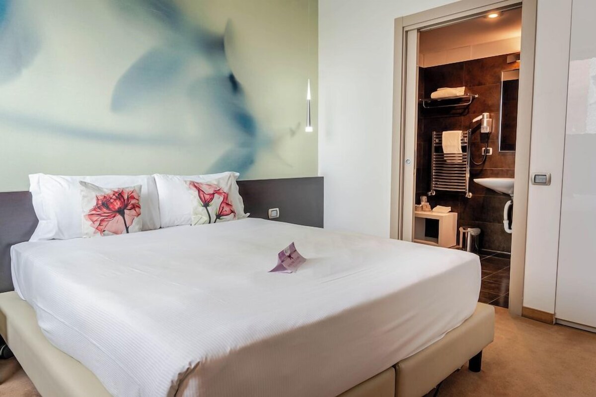 A hotel room with a bed, a bathroom with a shower, and a wall with a floral pattern.
