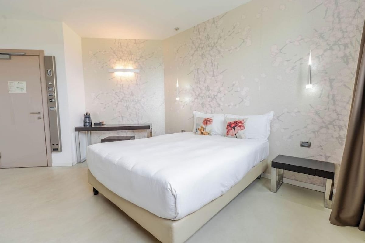 A hotel room with a bed, bedside table, desk, and a wall-mounted light fixture.