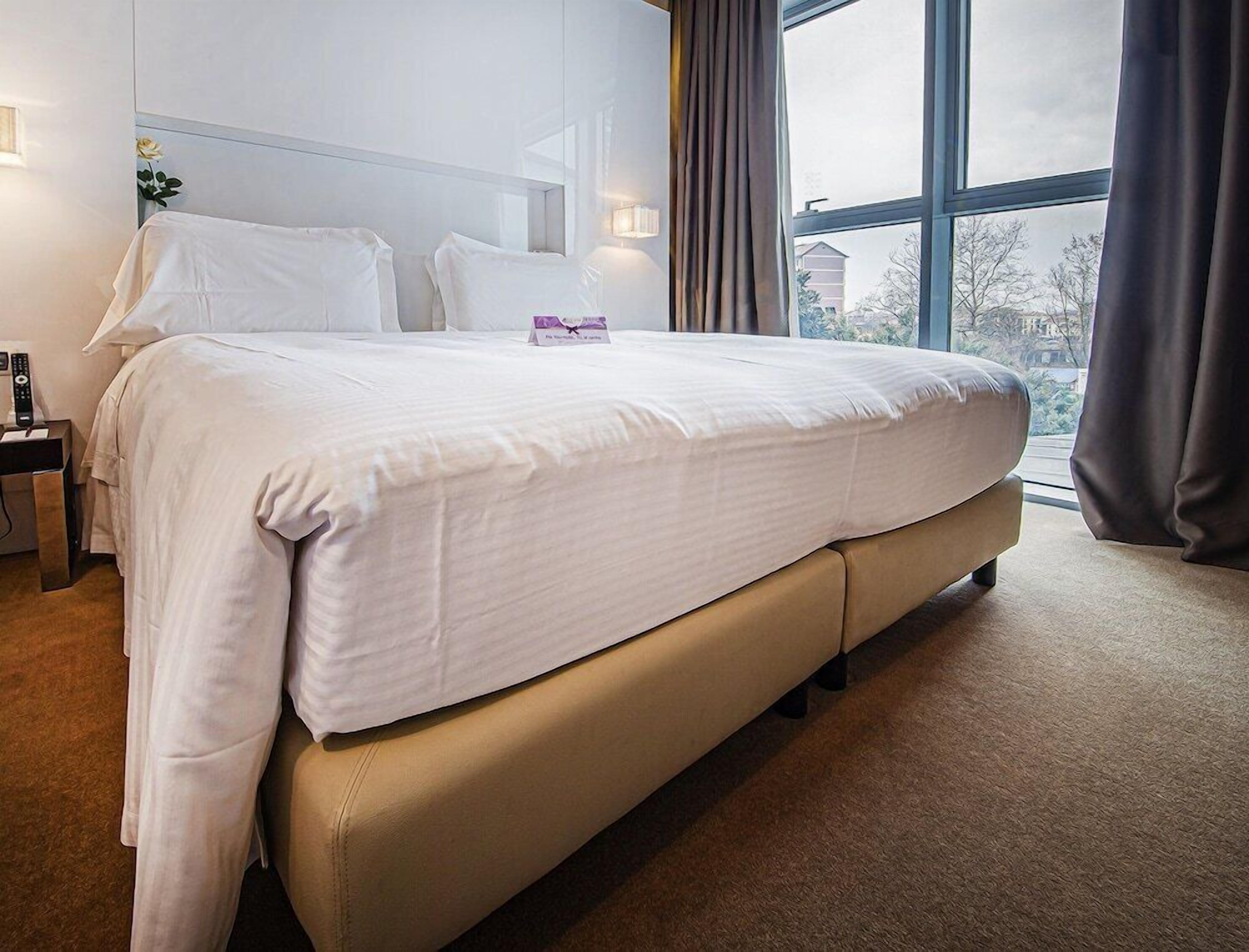 A neatly made bed in a hotel room with a large window, a bedside table with a lamp, and a remote control on the nightstand.