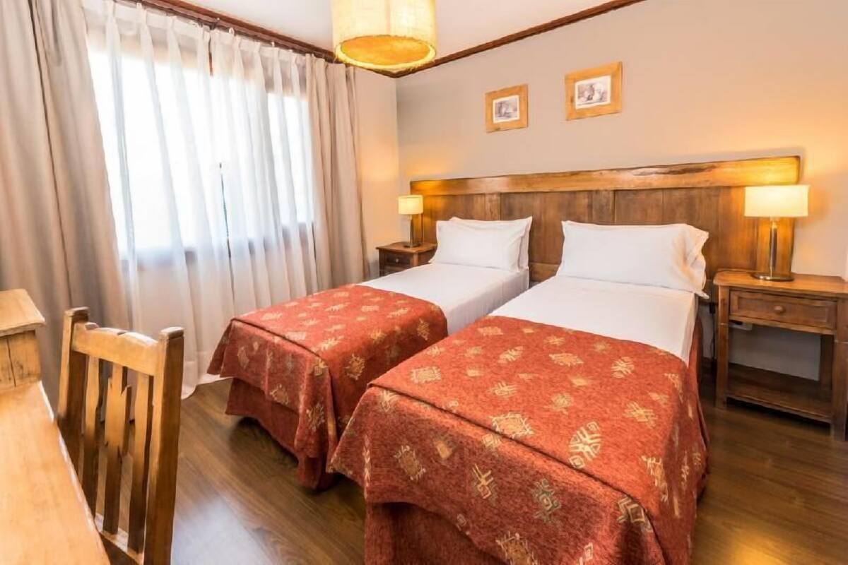 A hotel room with two beds, wooden headboard, a wooden chair, a desk, and a lamp.