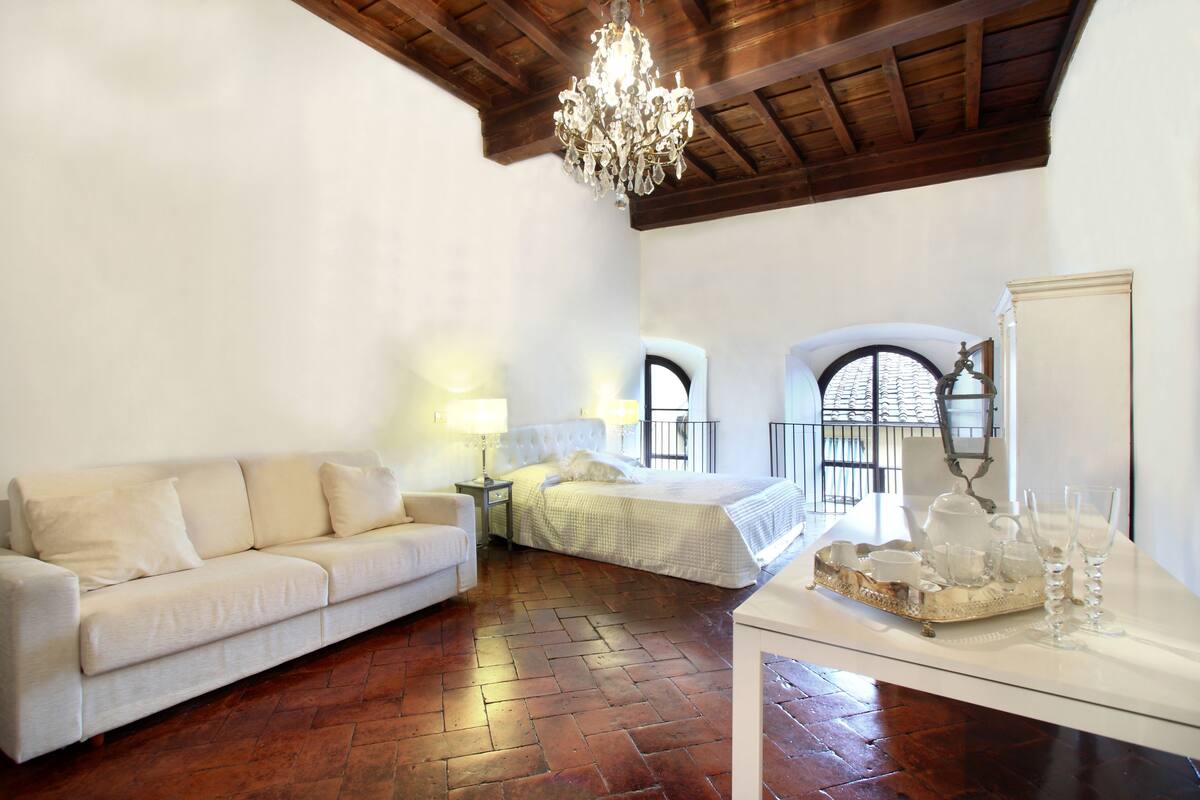 A room with a white sofa, a bed, a chandelier, and a table with glassware.
