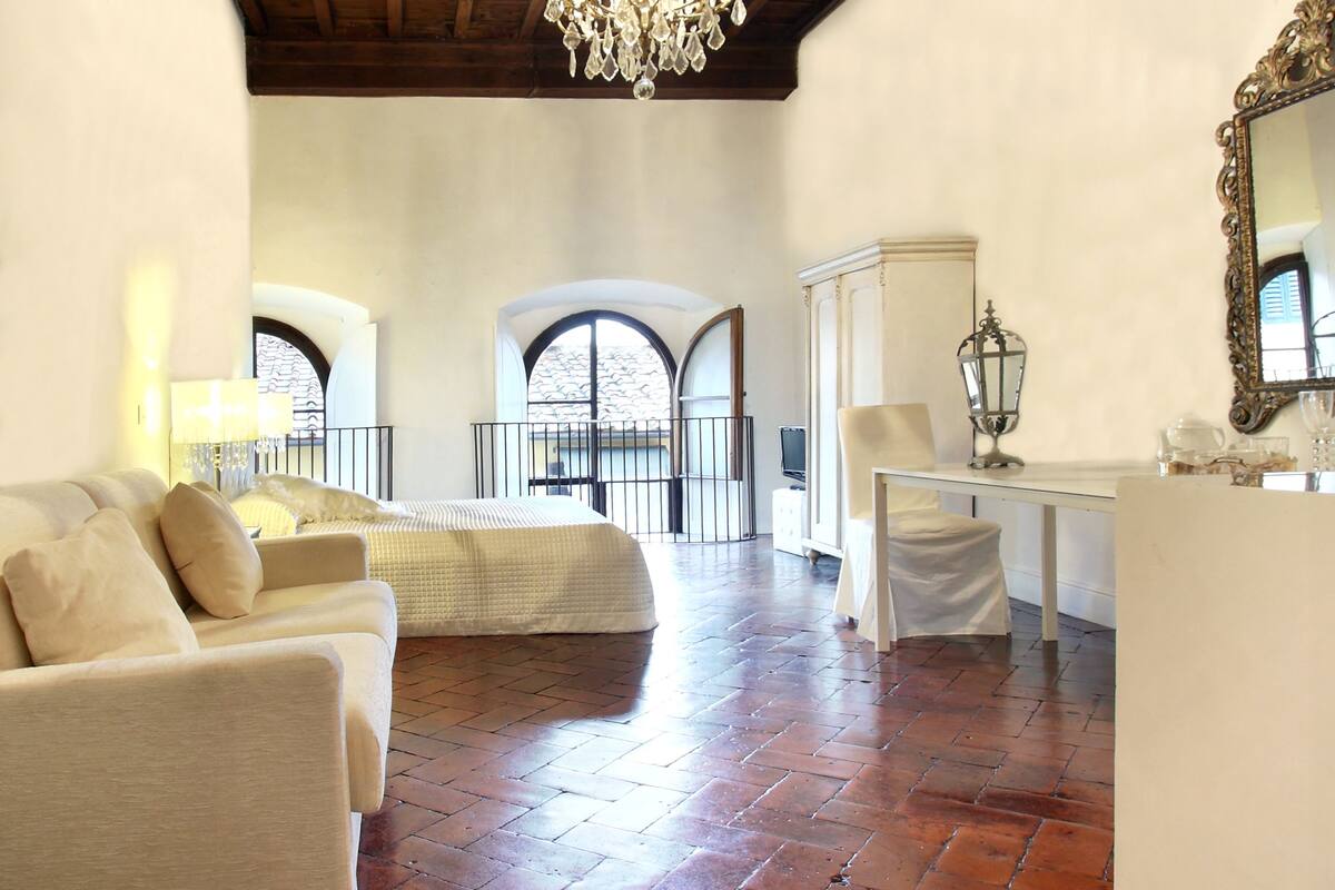 A spacious room with a high wooden ceiling, a chandelier, a white sofa, a dining table, and a view through arched windows.
