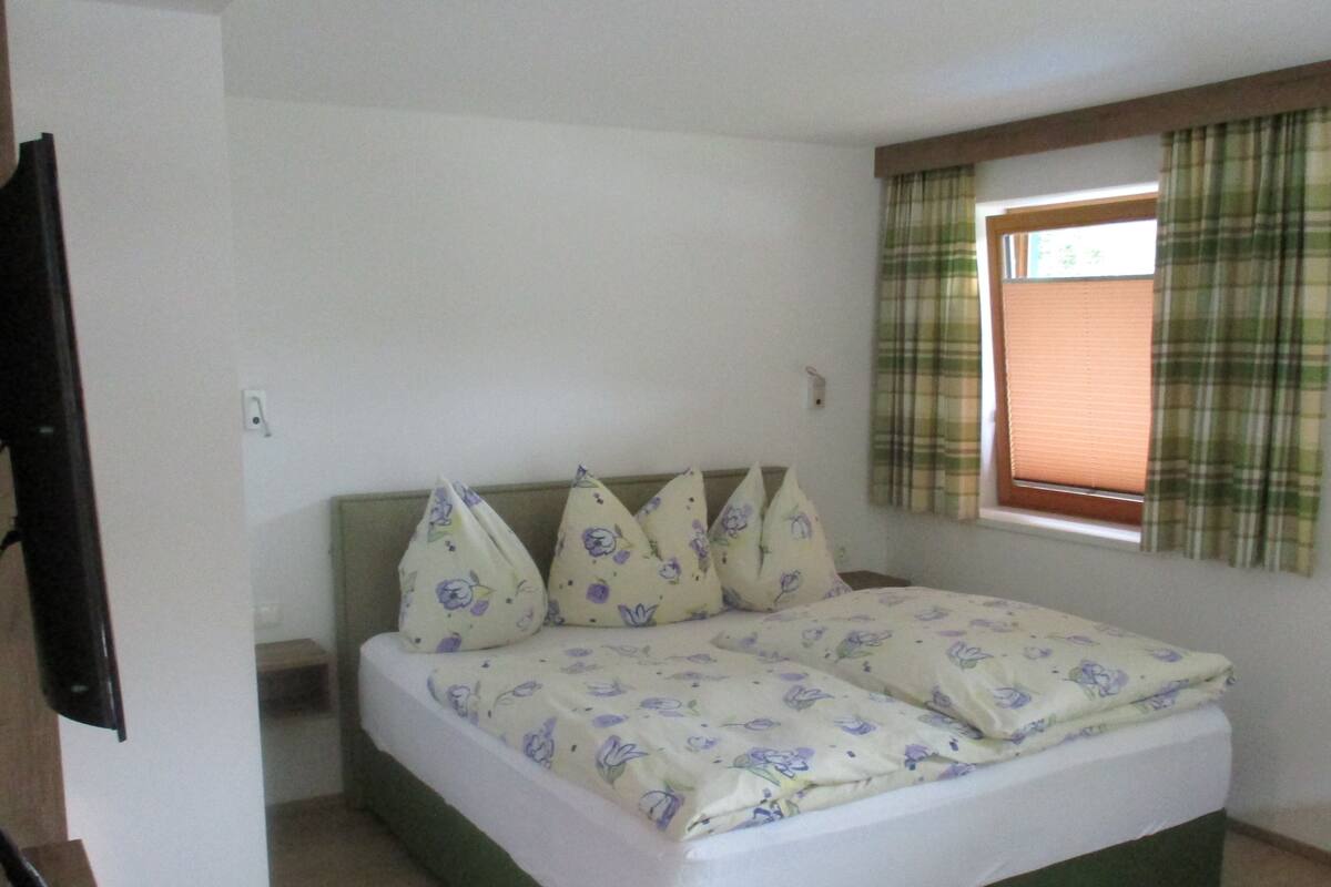 Ein Zimmer mit einem Bett, einem Vorhangfenster und einem Fernseher links.