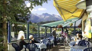 Un café en plein air avec des tables et des chaises, une vue sur la montagne et un espace abrité avec un auvent jaune.