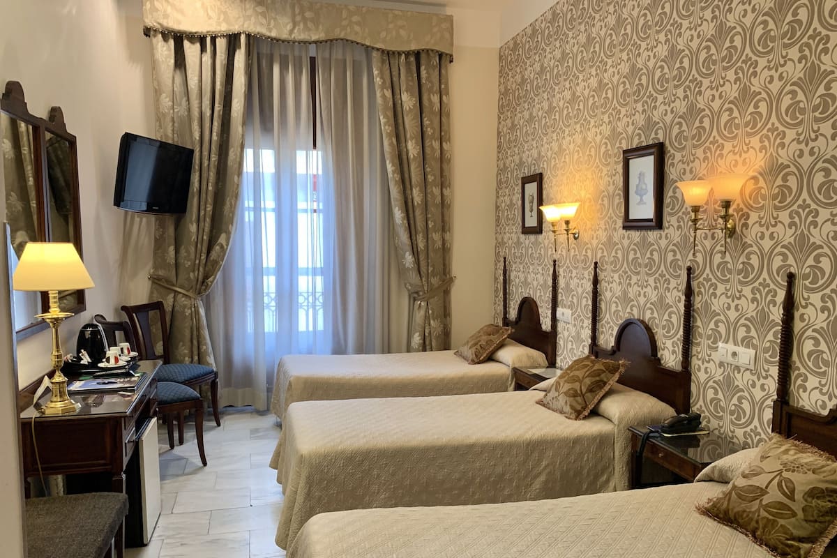 A hotel room with two beds, a desk with a lamp, a chair, a TV, and patterned wallpaper.