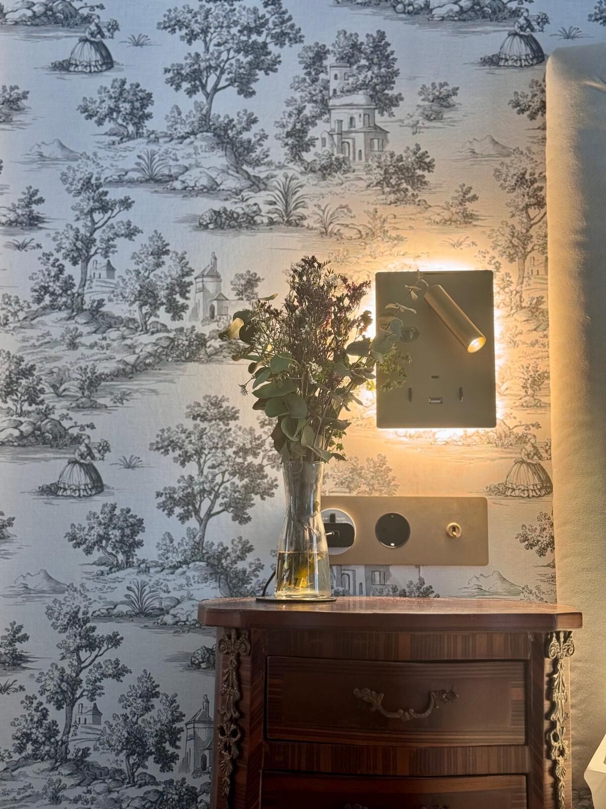 Una pared con diseño de papel tapiz antiguo, un jarrón con flores y una lámpara de pared.