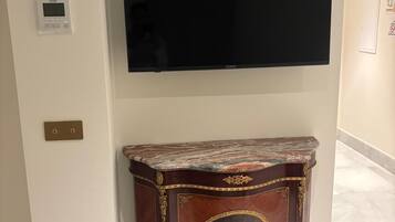 Una habitación con una televisión de pantalla plana montada en la pared, un mueble de tocador con cubierta de mármol y detalles dorados ornamentales, y un termostato en la pared.