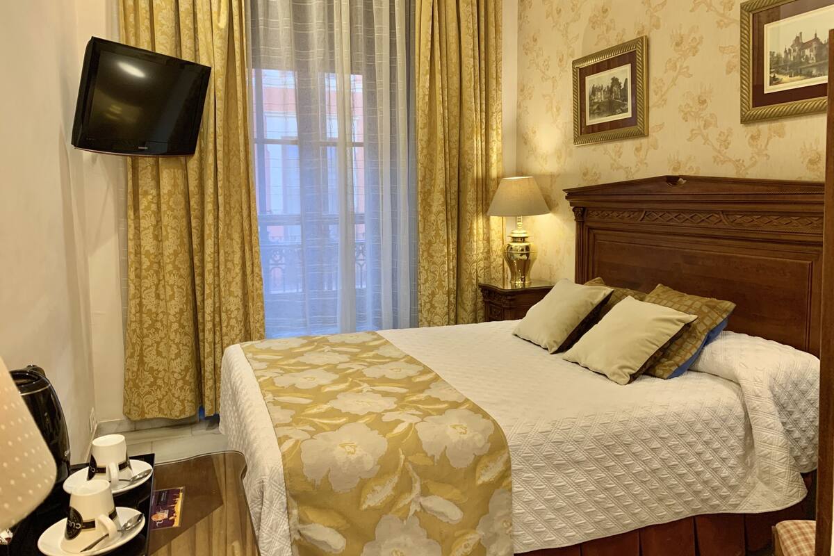 A hotel room with a bed, a TV mounted on the wall, a window with curtains, and two framed pictures on the wall.
