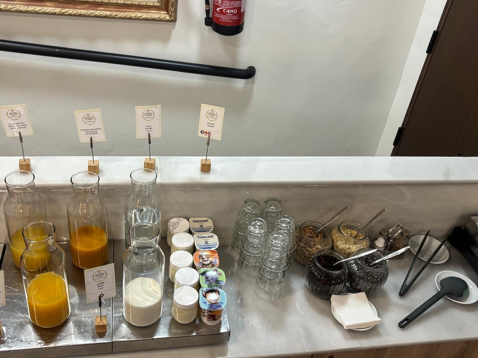 Een drankstation met verschillende drankjes en bekers, waaronder melk en sap, en een brandblusser aan de muur.